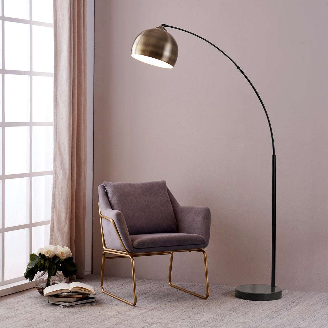 A Teamson Home Arquer Arc 68" Metal Floor Lamp with Bell Shade, Antique Brass in a room with a chair next to it.