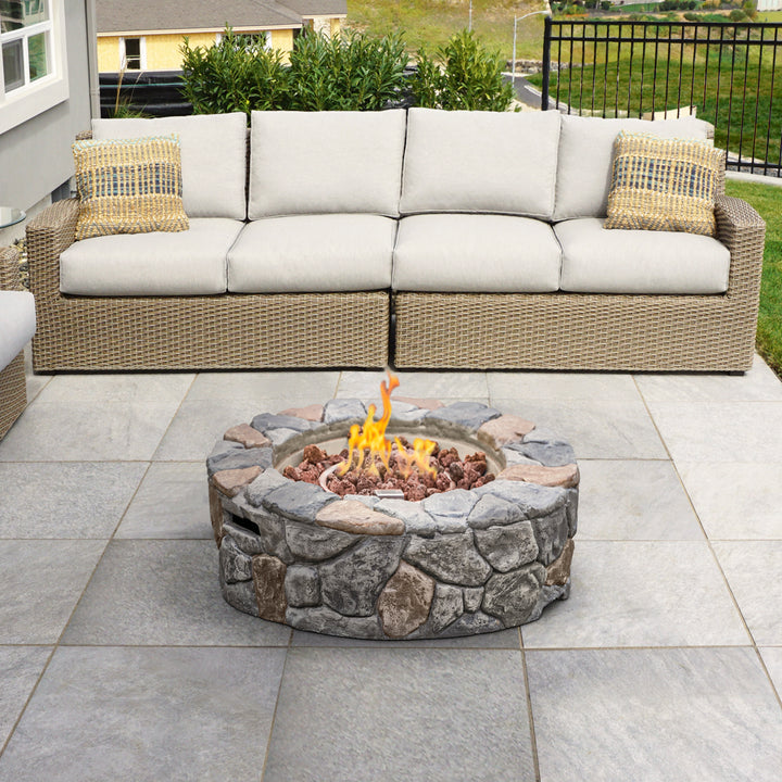 A modern outdoor living space featuring a Teamson Home 28" Outdoor Round Stone Propane Gas Fire Pit in Stone Gray and a sectional wicker sofa with plush cushions and throw pillows.