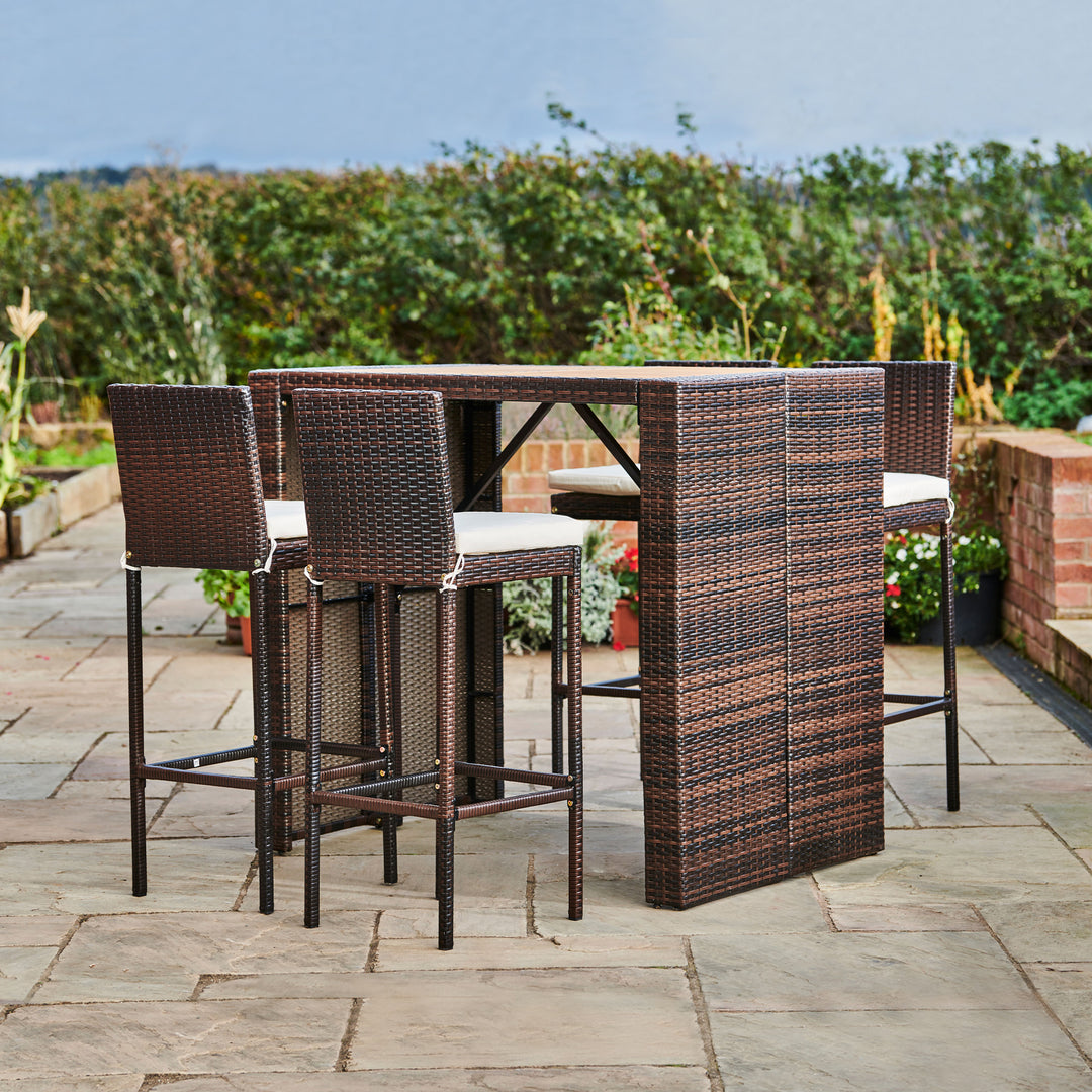 A Teamson Home Outdoor Brown PE Rattan & Acacia 5-Piece High Top Table & Stools with White Cushions with tall shrubbery in the background
