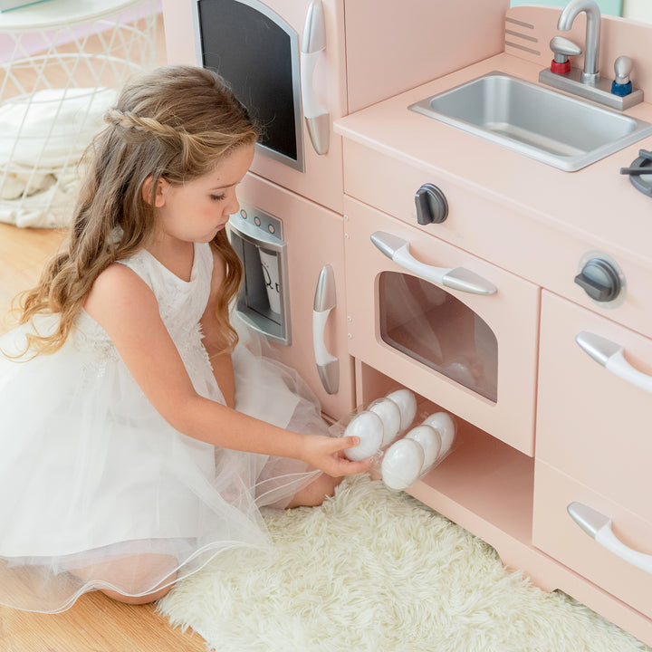 A young girl in a white dress playing with a Teamson Kids Little Chef Fairfield Retro Kids Kitchen Playset with Refrigerator, Pink.