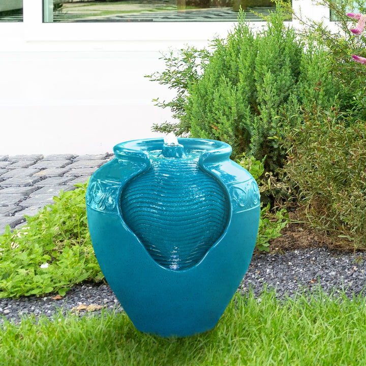 A teal Teamson Home Outdoor Glazed Pot Floor Fountain with LED Lights, positioned on a garden path beside green shrubbery, serves as both practical garden decor and a durable lawn statue.