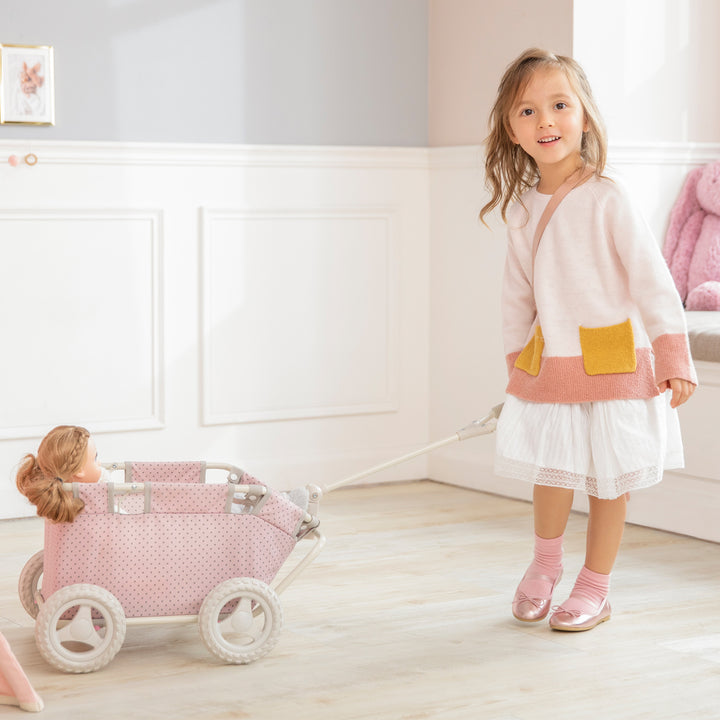 A little girl is pushing a pink doll in an Olivia's Little World Polka Dots Princess Baby Doll Wagon, Pink adorned with polka dots.
