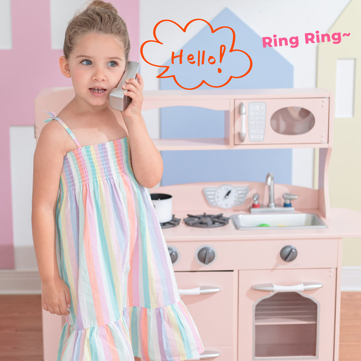 A young child pretending to talk on a toy telephone in front of the Teamson Kids Little Chef Westchester Retro Play Kitchen, Pink.
