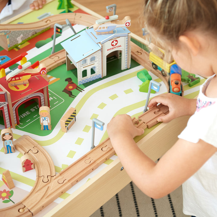 A little girl connecting pieces of the train track together on top of the fully-illustrated table.