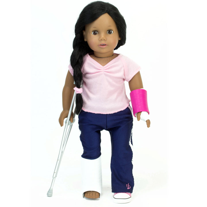 A black-haired 18" doll with a cast on her right foot, a cast and wrap on her left arm, standing against a single crutch on her left side.