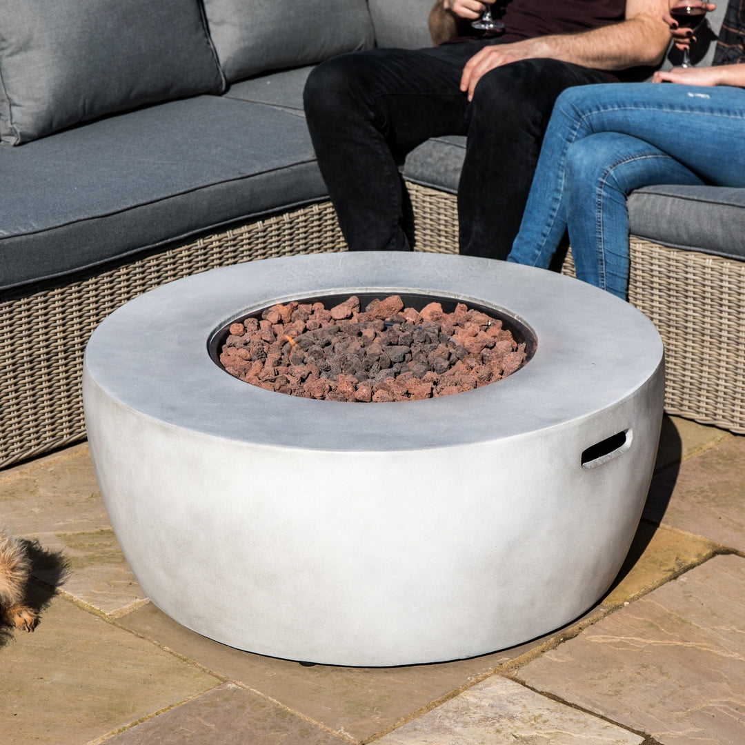 Teamson Home 36" Outdoor Round Propane Gas Fire Pit with Faux Concrete Base, Gray on a patio next to a wicker sofa with people seated nearby.
