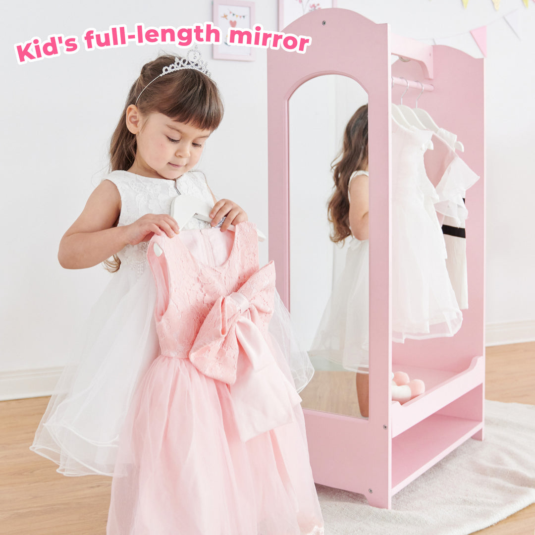 A girl in a white dress holding a pink dress in front of the mirror of a pink wardrobe with some white dresses hanging from it and a caption "Kids full-length mirror"