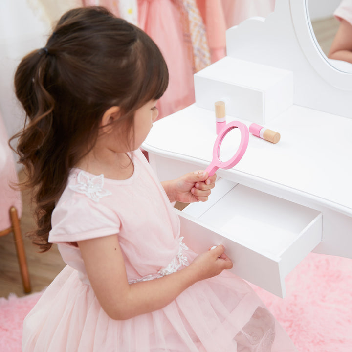 A little girl sitting at the white vanity table looking at herself in a pink hand held mirror.