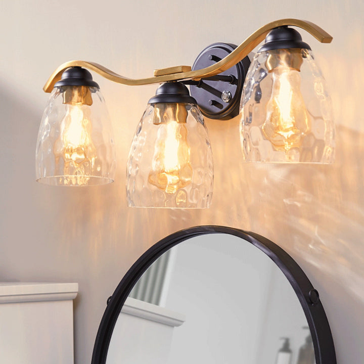 Heidi 3-Light Bathroom Vanity Wall Sconce Light with 3-Stage Touch Dimmer & Clear Hammered Glass Cloche Shades, Black/Brass features three illuminated clear hammered glass cloche shades above a round mirror.