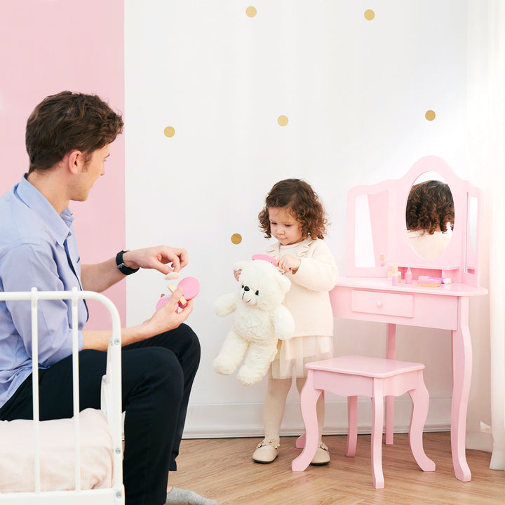 A little girl brushing a stuffed polar bear next to a pink vanity table and stool with a tri-fold mirror with her dad sitting on a bed.