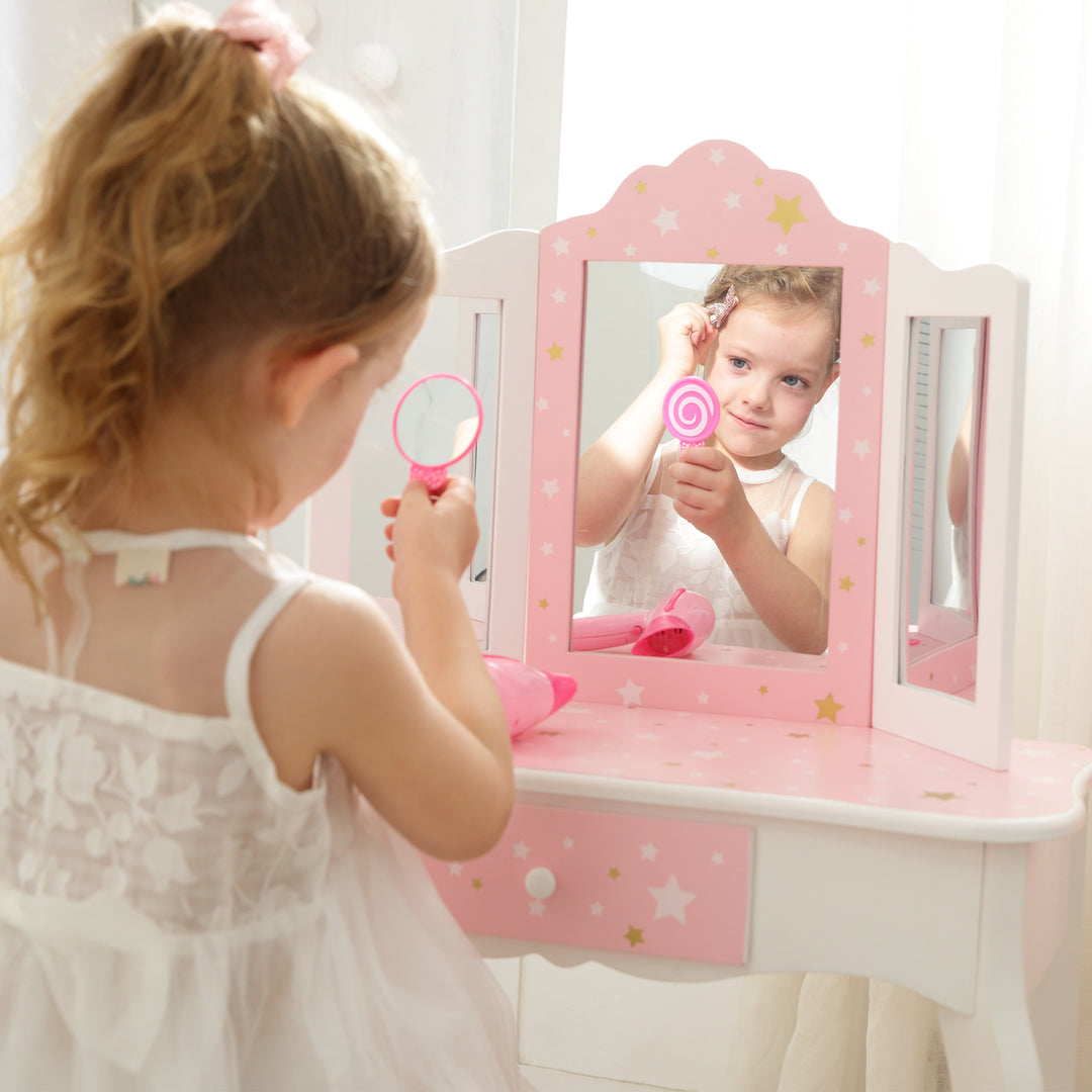 A little girl in a pink dress looking at a Fantasy Fields Gisele Play Vanity Set with Mirrors, Pink/White.