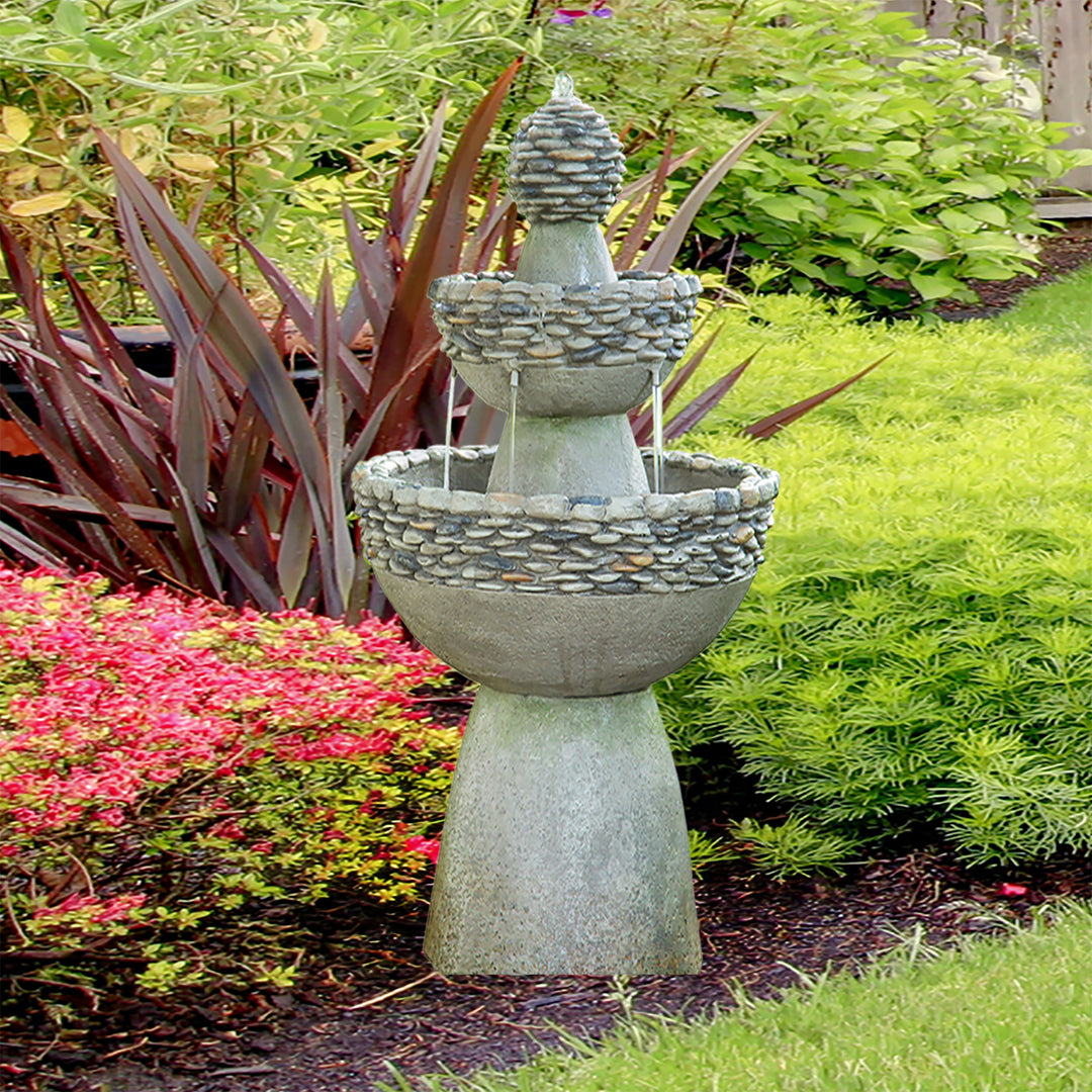 A Teamson Home Outdoor 3-Tier Pedestal Floor Fountain, Gray, set amongst some flowering bushes and decorative grasses