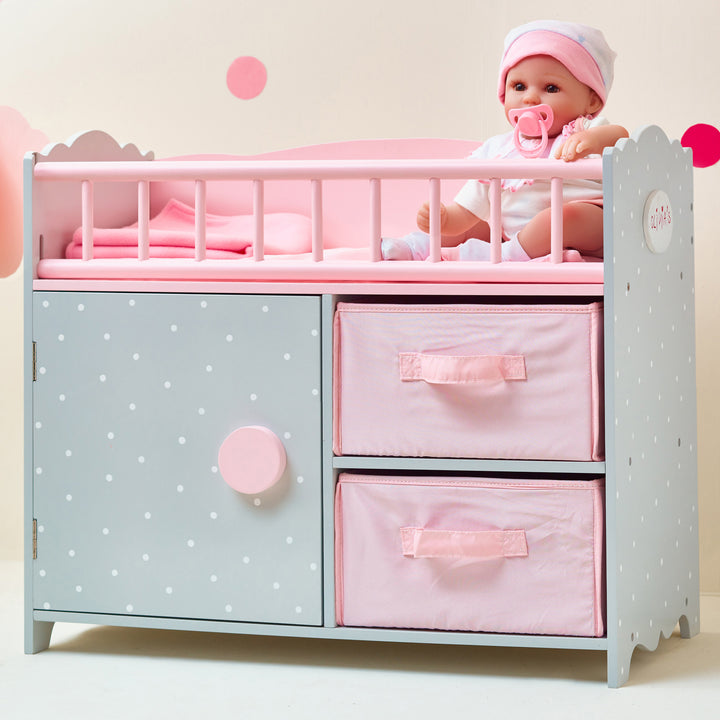 A baby doll sitting in the crib portion of a gray baby doll crib with white polka dots and pink accents.