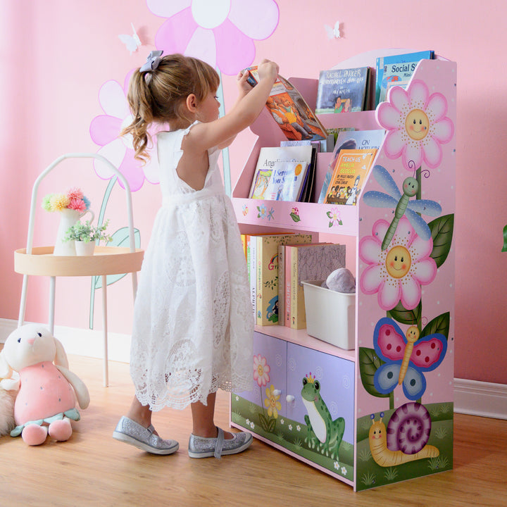 A girl in a white dress standing next to a Fantasy Fields Magic Garden Kids 3-Tier Wooden Bookshelf with Storage, Multicolor.