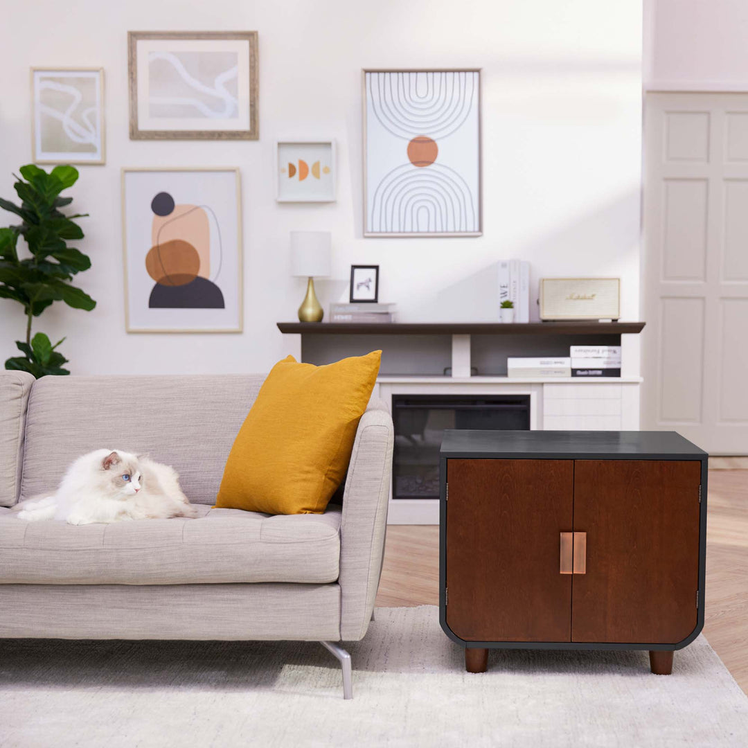 Teamson Pets Small Dyad Wooden Cat Litter Box Enclosure and Side Table, Mocha Walnut, next to a flax-colored sofa with a white cat sitting on the sofa.