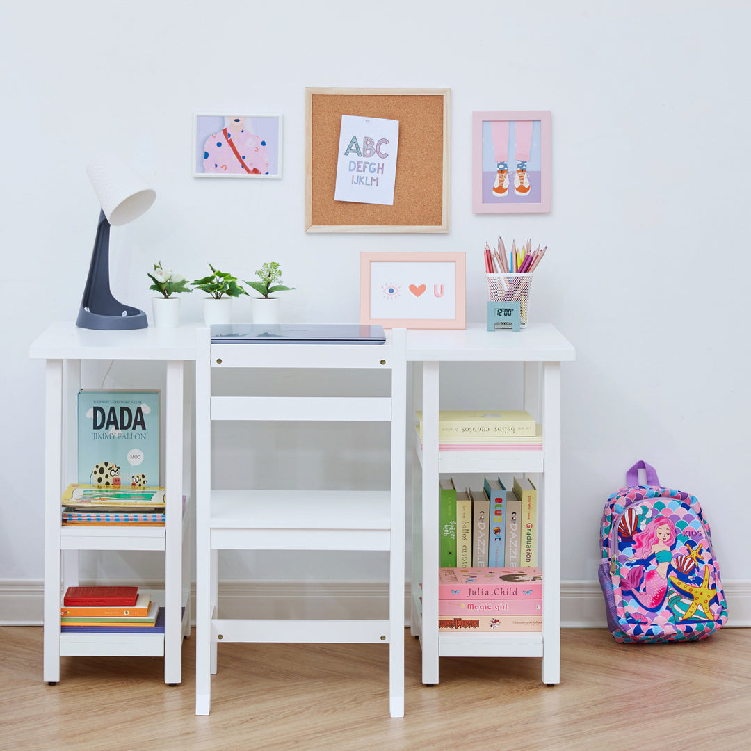 A children's white desk and matching chair against a white wall with books on the shelves, laptop and desklamp, and framed pictures on the wall.