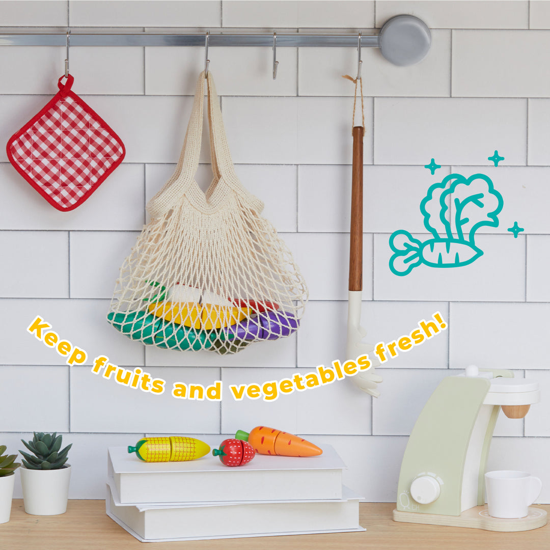 A bright Teamson Kids Little Chef Frankfurt 21 Piece Wooden Produce Shopping Bag setting with a hanging net bag full of colorful pretend food, a quirky drawing of vegetables on the wall, and a modern food slicer on the counter, all designed with kid-sized dimensions.