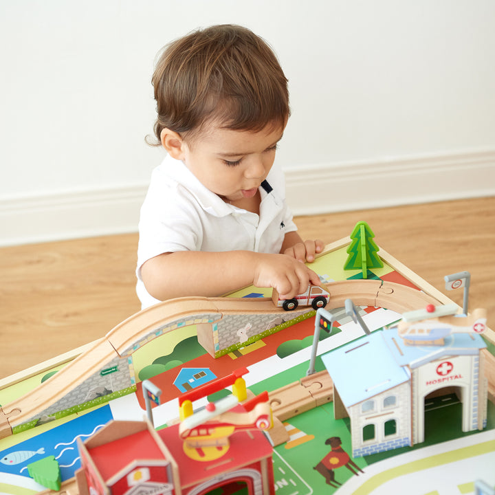 A young child pushing a white car down the tracks on top of the train table past a pretend table and behind a set of buildings.