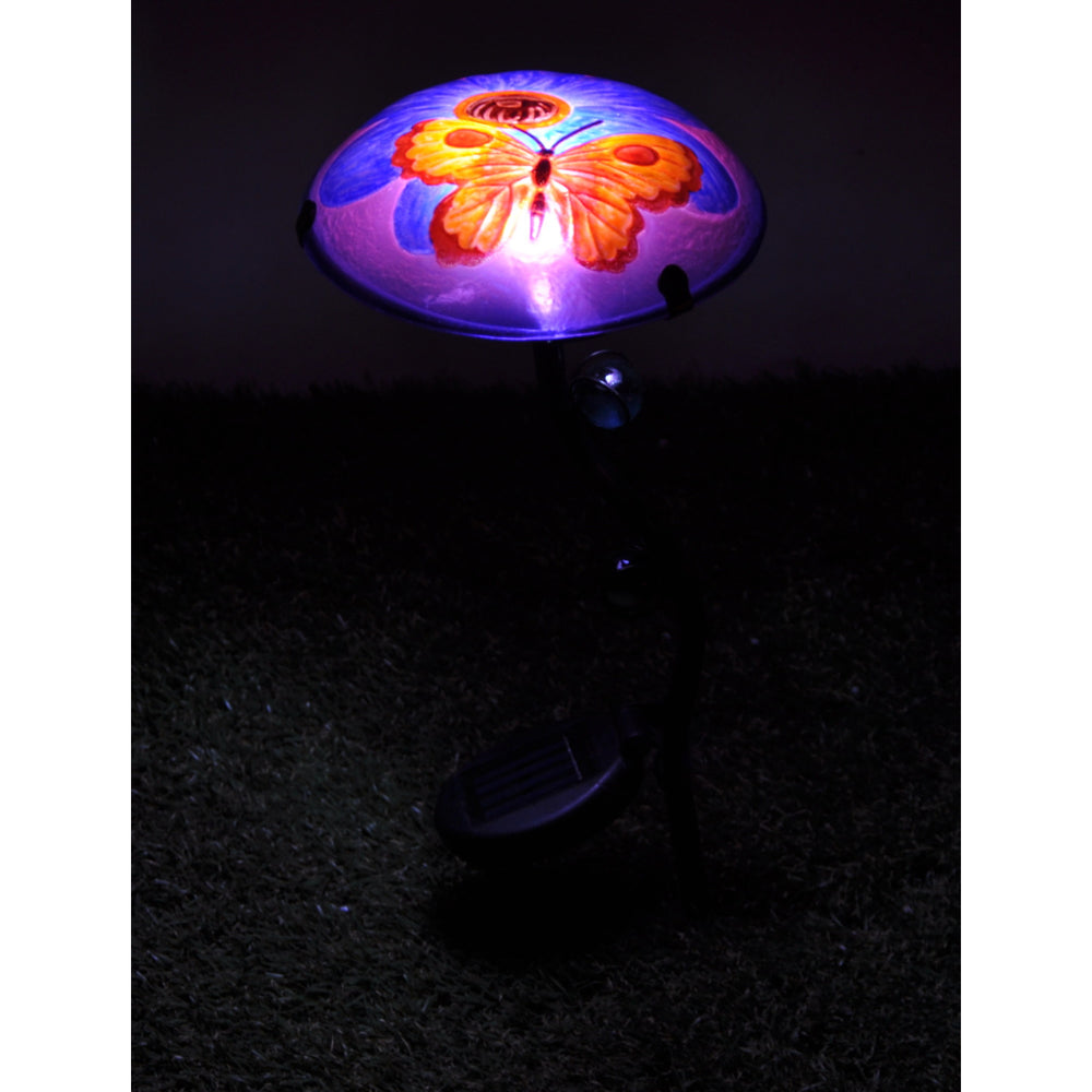 A Teamson Home Outdoor Butterfly Fusion Glass Solar Powered LED Light Stake, Purple with a butterfly design, featuring a fusion glass solar panel, stands on a garden stake glowing at night.