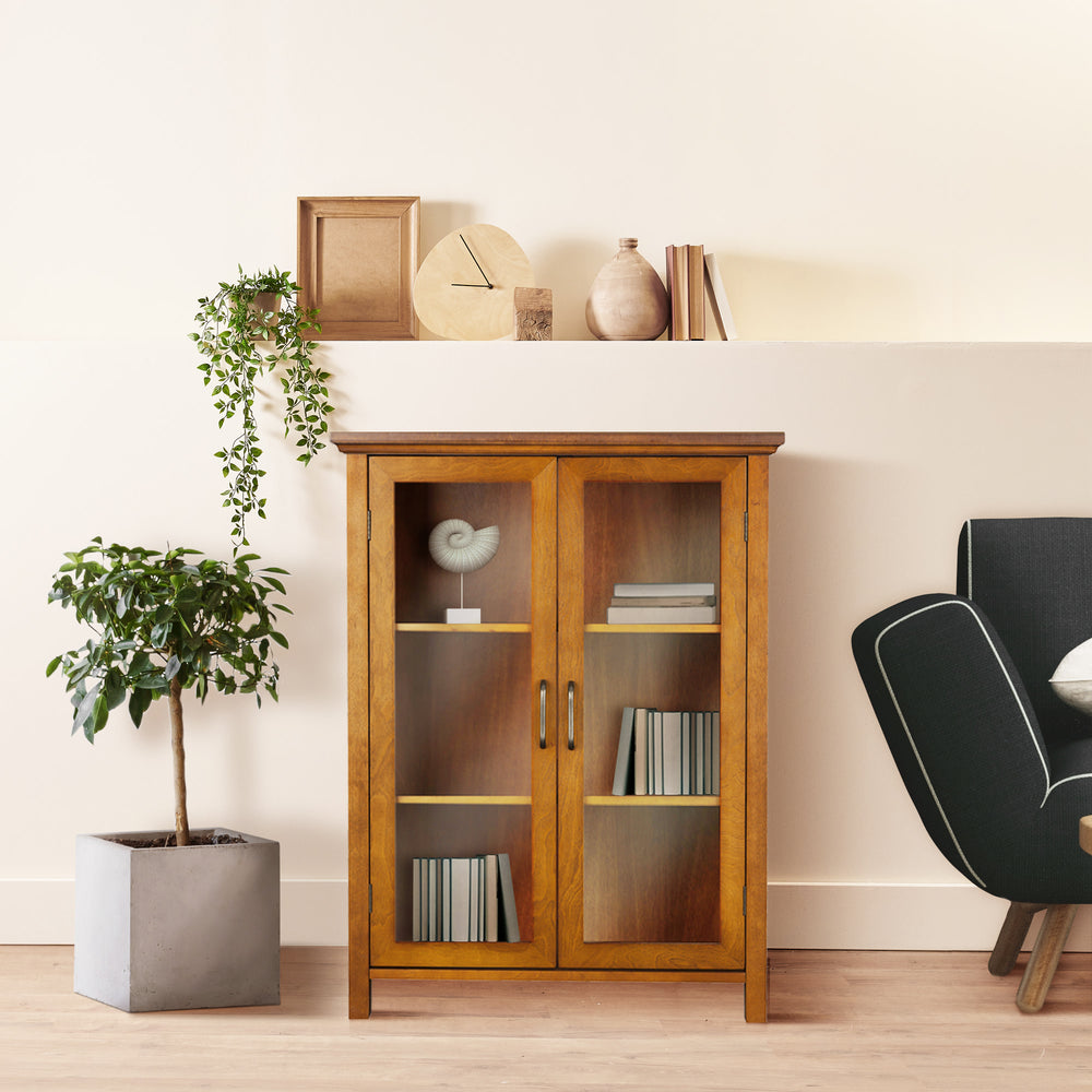 A Teamson Home Avery Wooden 2 Door Floor Cabinet with Storage, Oiled Oak containing books and decorative items, flanked by a potted plant on one side and a black armchair on the other against a neutral wall.