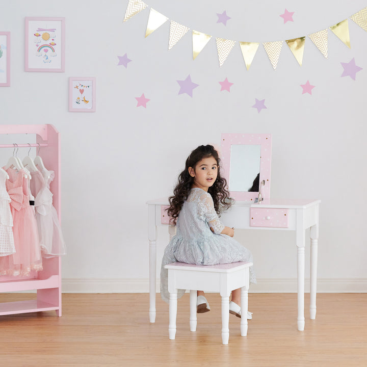 A little girl sitting in front of a pink Fantasy Fields Kids Kate Twinkle Star Vanity Set with Foldable Mirror and Chair.