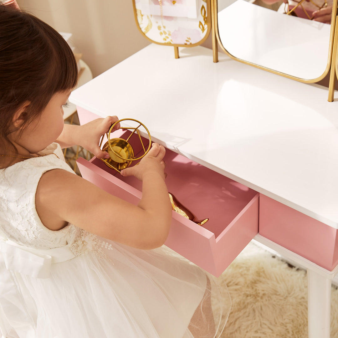 A little girl putting a gold cup in a pink drawer of her pink and white vanity set.