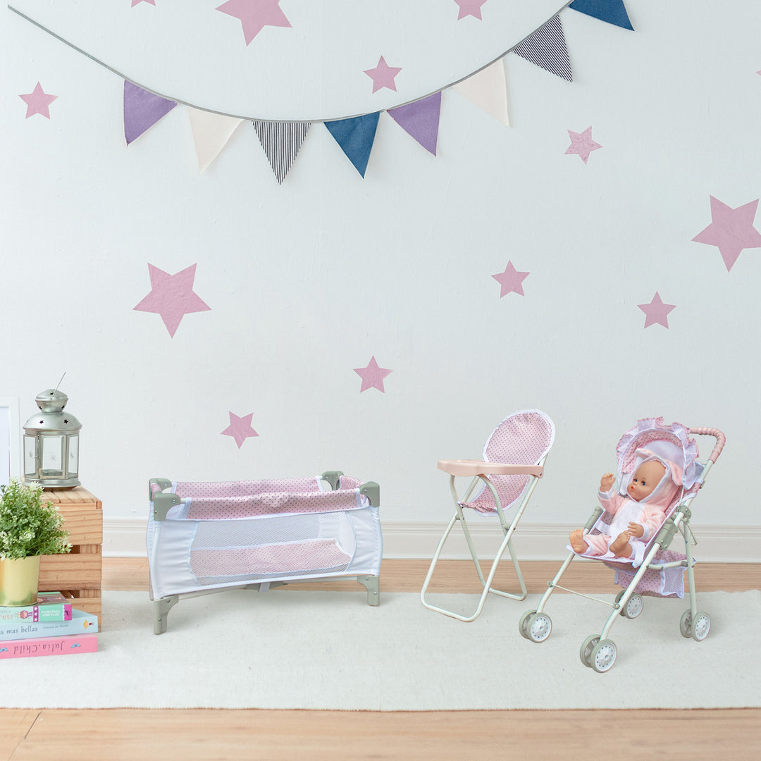A playroom with pink stars on a white wall and a white and pink doll play pen, doll high chair, and a doll stroller (with a doll sitting in it) in front.
