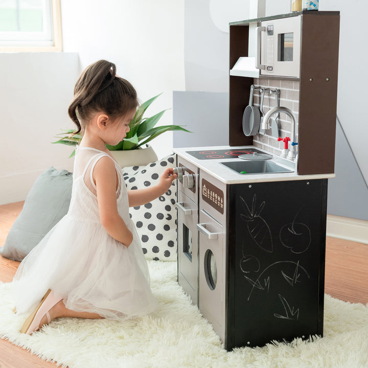 A young girl playing with a Teamson Kids Little Chef Burgundy Classic Play Kitchen with a chalkboard panel on the side.
