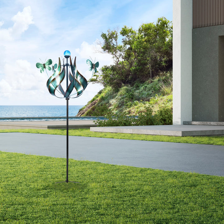 A whimsical Teamson Home Outdoor Solar Tulip and Butterfly Kinetic Windmill Sculpture with LED Light, Teal, now a solar-powered light, stands near a coastal walkway with the ocean in the background.