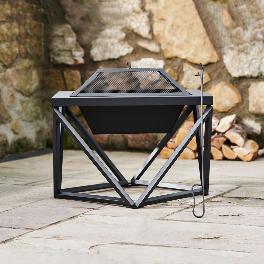 A Teamson Home Outdoor 24" Wood Burning Fire Pit with Tabletop and Decorative Base, Black on a slate patio surface with a spark screen and poker