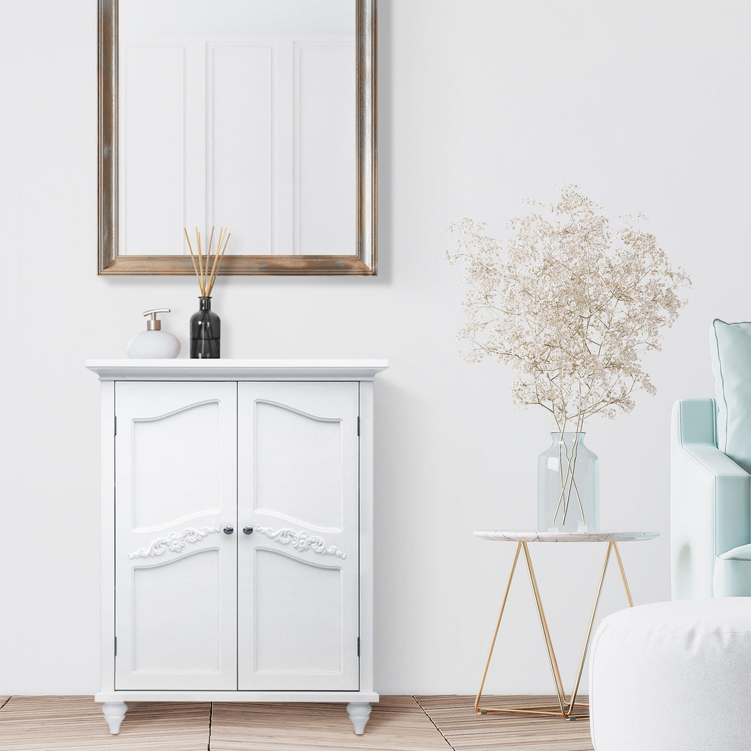 A modern bathroom corner featuring a Teamson Home Versailles Wooden Floor Cabinet White, decorative plant, side table, and a framed mirror on a clean white wall.