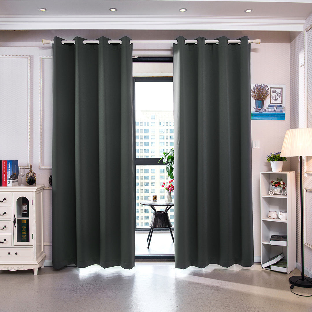 A brightly lit room with a pair of Teamson Home 96" Delphi Premium Solid Insulated Thermal Blackout Window Curtain Panels with Grommets, Smoke Gray drawn across a window, with a bookshelf and a lamp to the side.