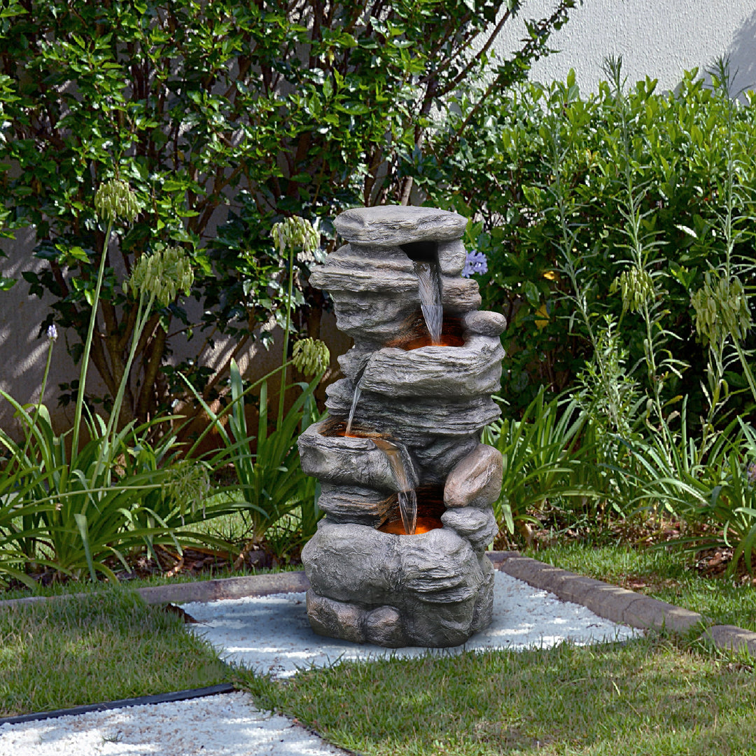 Teamson Home 4-Tier Stacked Stone Water Fountain with LED Lights, Gray, set next to some shrubs in a back yard
