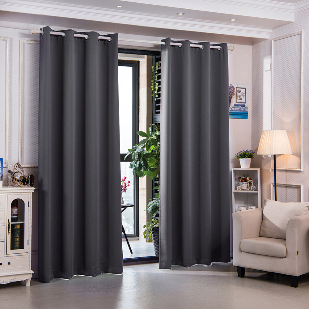 A well-lit modern living room with dark Teamson Home 84" Sparta Premium Solid Insulated Thermal Blackout Window Curtain Panels with Grommets, Dove Gray, a cream armchair, and decorative plants near an open door leading to a balcony.