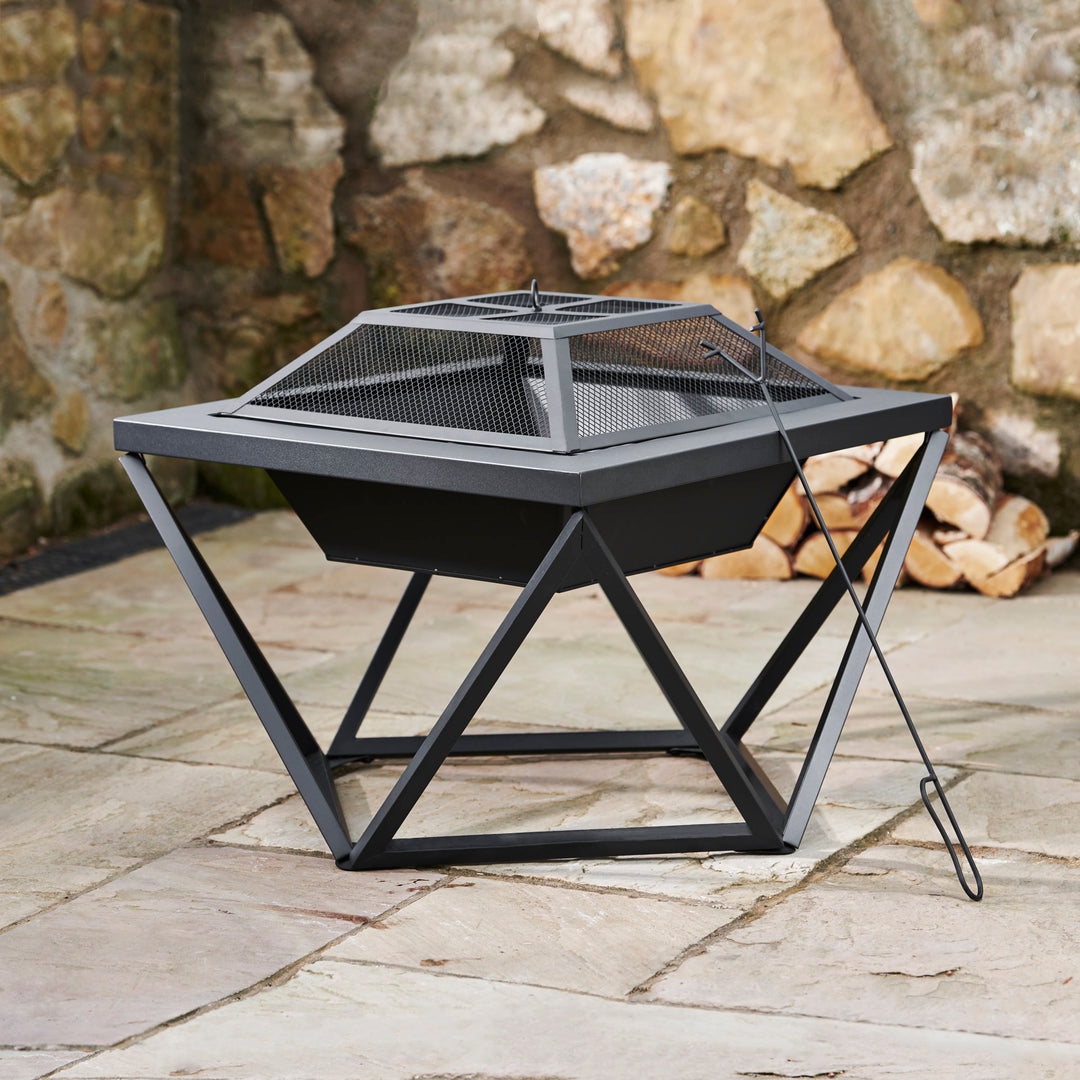 A Teamson Home Outdoor 24" Wood Burning Fire Pit with Tabletop and Decorative Base, Black, with a spark screen and poker on a slate patio surface