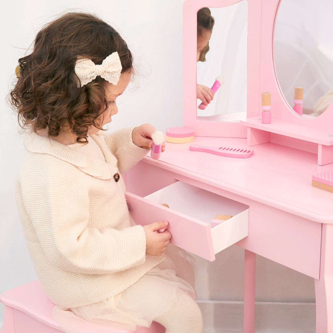 A little girl opening the drawer of a pink vanity table and stool with a tri-fold mirror.