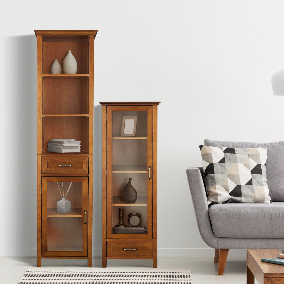 A well-organized living room corner featuring a Teamson Home Avery Wooden Linen Tower Cabinet with Storage, Oiled Oak next to a gray couch with a patterned cushion.