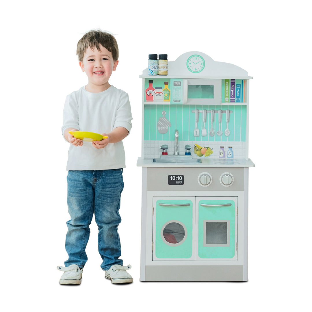 A smiling child standing next to a Teamson Kids Little Chef Madrid Classic Play Kitchen with Salt & Pepper Shakers, Mint/Gray, holding a yellow plate.