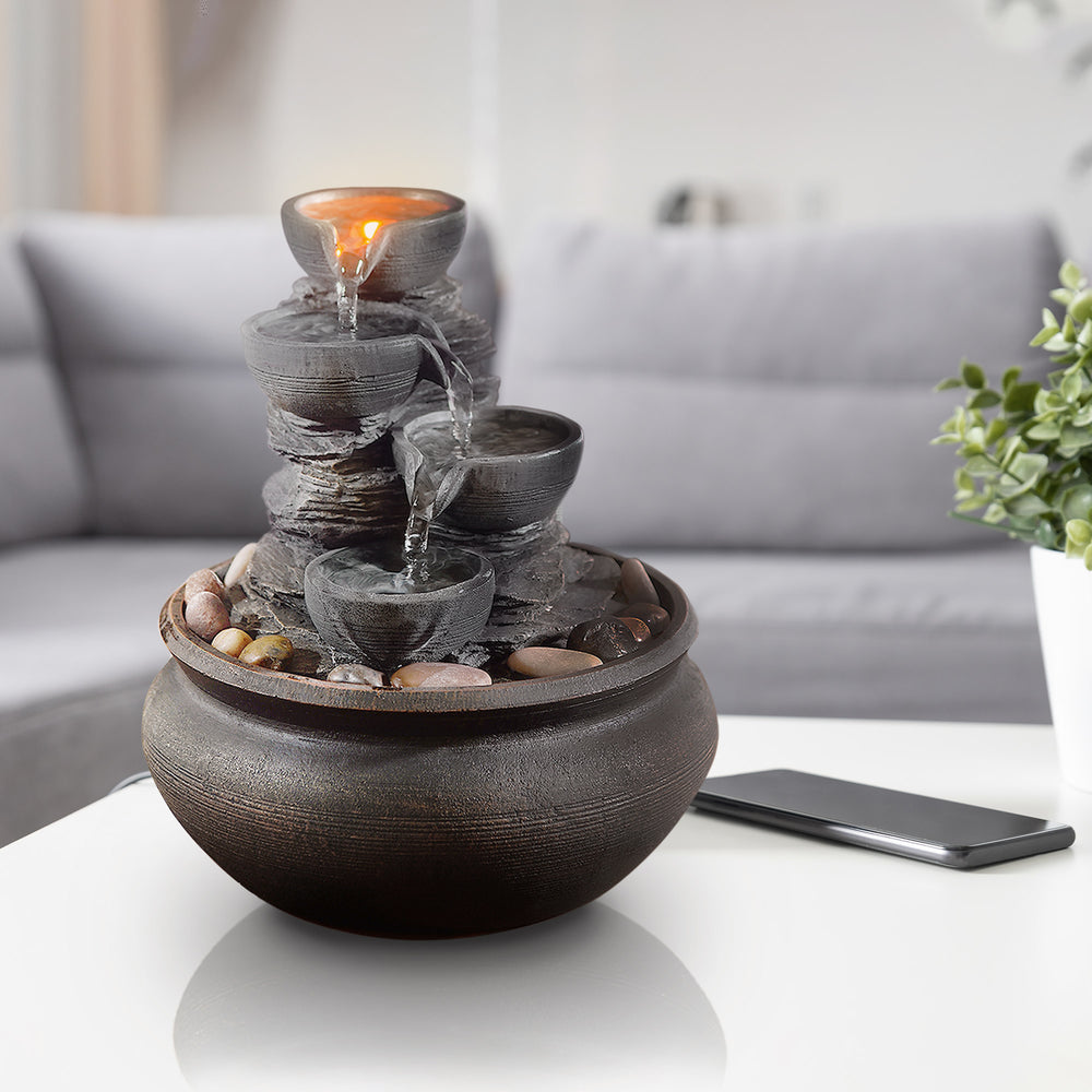 Teamson Home Tabletop Water Fountain, Stone Gray, with four tiers and pebbles on the base, placed on a table in a modern living room setting.