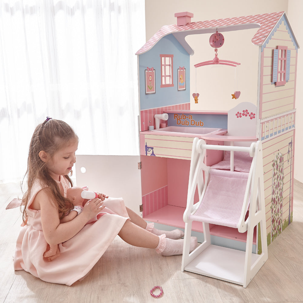 A little girl holding her baby doll sitting next to a dollhouse/baby doll nursery playset with an individual baby doll swing.