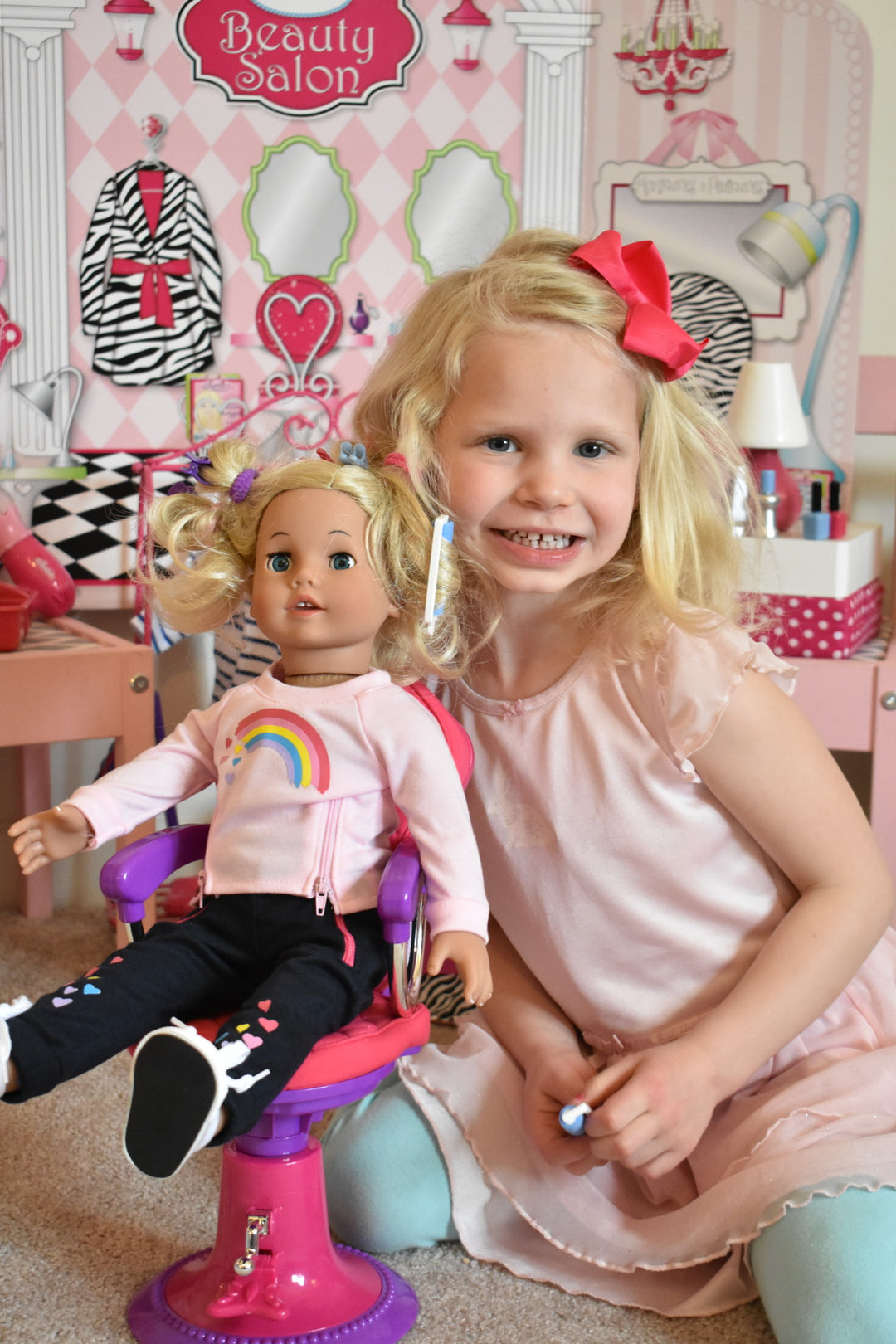 A little blonde girl sits next to her blonde doll playing beauty salon.