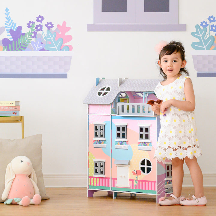 A little girl standing in front of a Teamson Kids Sunroom Dollhouse with 11 Accessories, Multicolor.