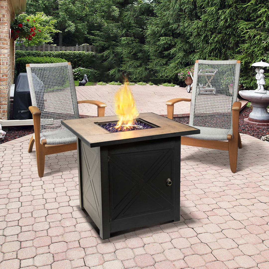 Two chairs surrounding a Teamson Home Outdoor Square 30" Propane Ceramic Gas Fire Pit with Steel Base, Black on a brick patio.