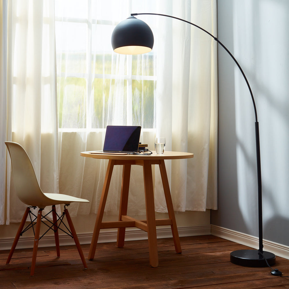 A table with a Teamson Home Arquer Arc Metal Floor Lamp with Bell Shade, Black and a chair in front of a window.