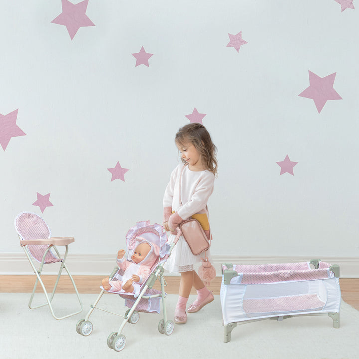 A little girl with pushing a baby doll in a pink and white stroller. There is a doll high chair on the left and a doll play pen on the right, both in pink and white.