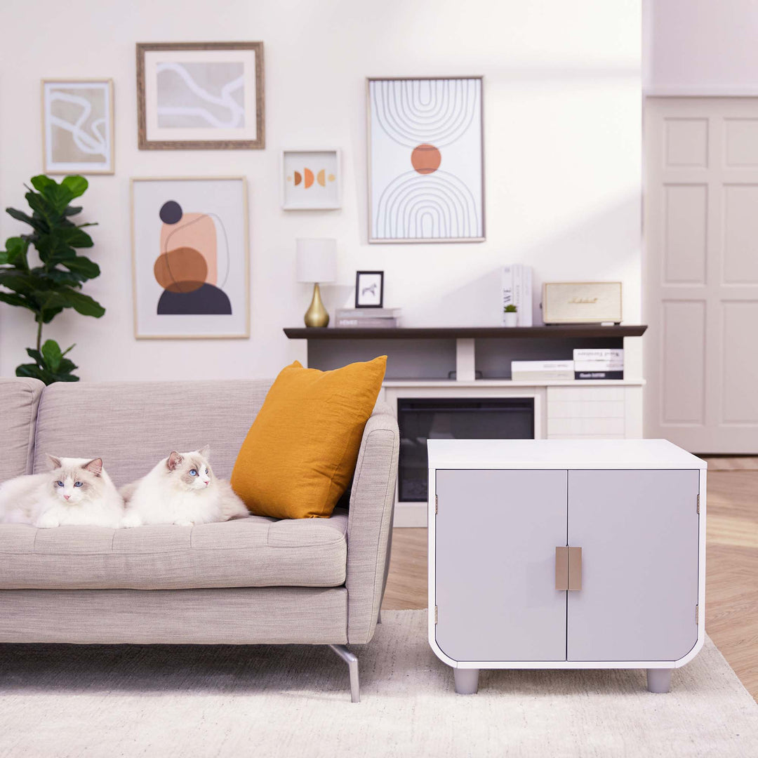 Teamson Pets Dyad Wooden Cat Litter Box Enclosure and Side Table, Alpine White, next to a flax-tone sofa with two cats on it.