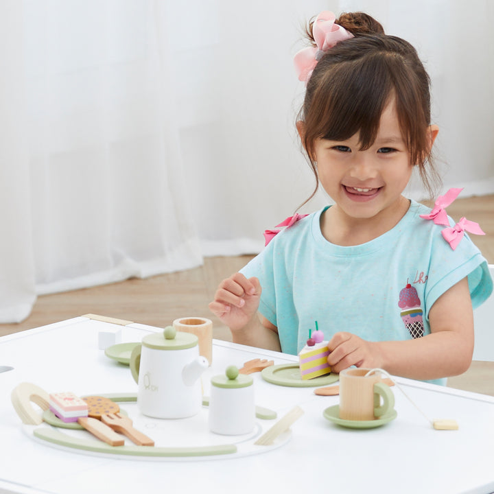 A young girl playing with a Teamson Kids Little Chef Frankfurt 20 Piece Wooden Play Kitchen Tea Party Set, Green, smiling brightly.