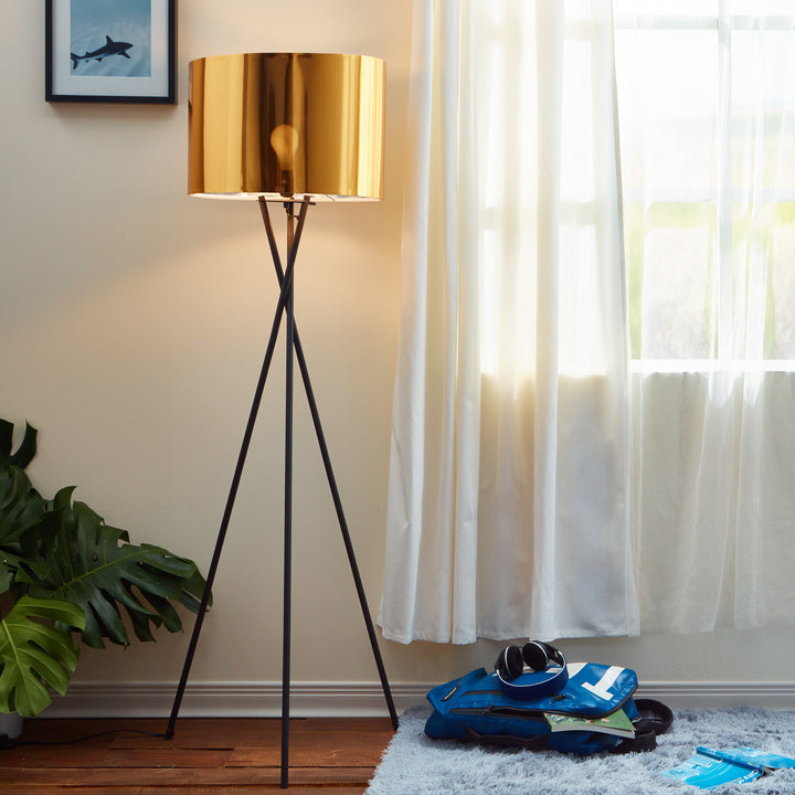 A Teamson Home Cara 62" Modern Tripod Floor Lamp with Metal Drum Shade, Black/Gold, n a living room next to a picture window