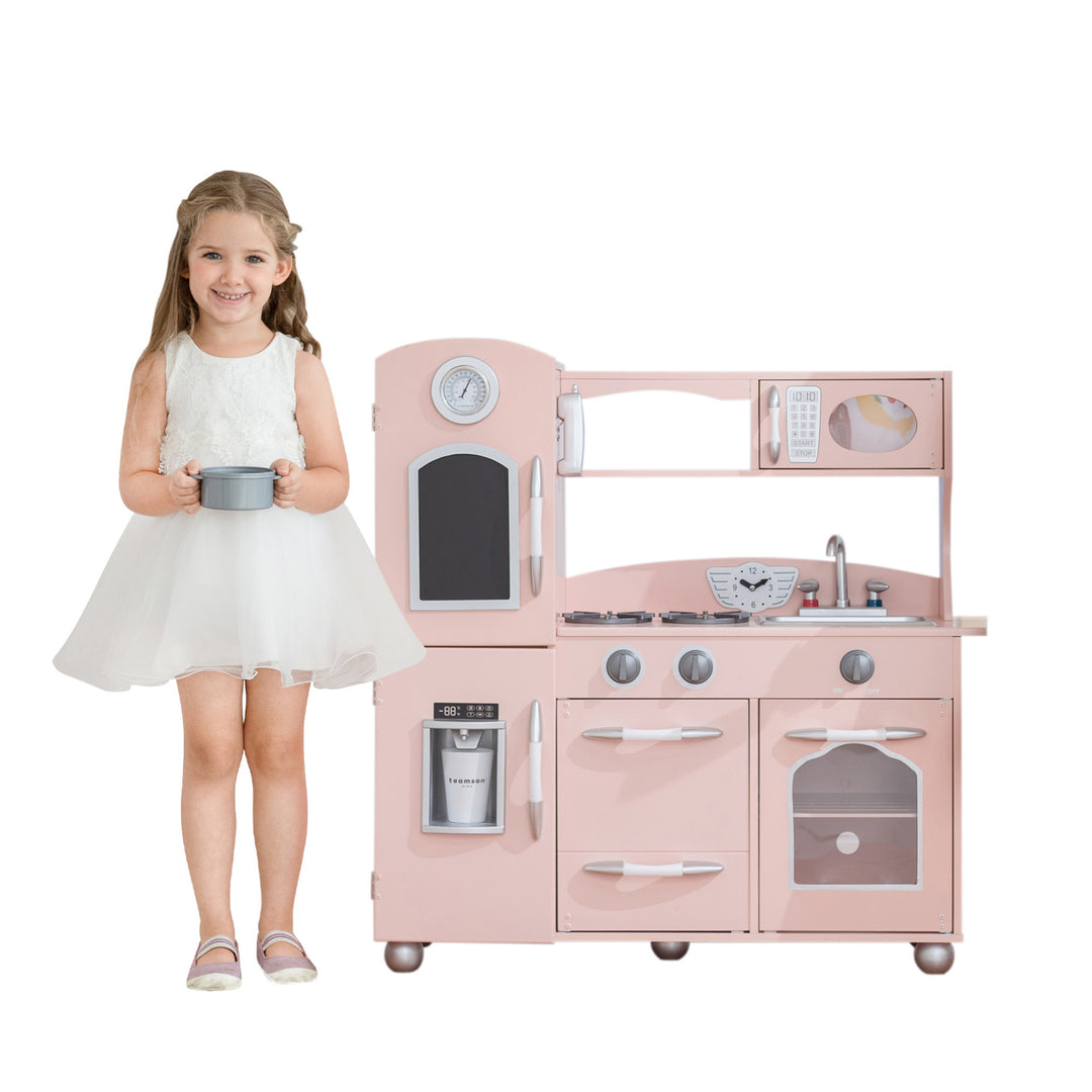 A young girl smiling and holding a pot in front of a Teamson Kids Little Chef Westchester Retro Play Kitchen, Pink with interactive features.