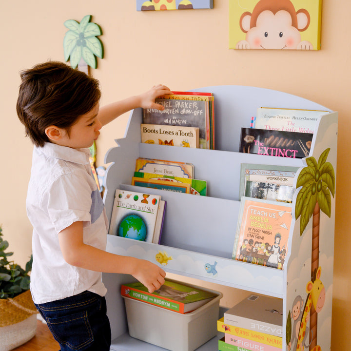 A boy is standing in front of a Fantasy Fields Sunny Safari Kids 3-Tier Wooden Bookshelf with Storage, Multicolor.